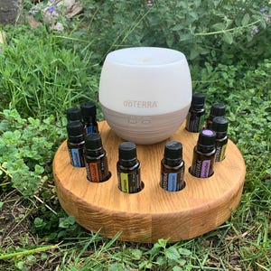 Peut inclure: Un diffuseur d'huiles essentielles blanc et beige avec le logo "dōTERRA", entouré de petites bouteilles d'huiles essentielles. Les bouteilles sont placées dans un support rond en bois. La scène se déroule à l'extérieur sur de l'herbe verte.