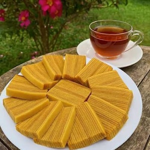 Puede incluir: Un plato blanco contiene varias rebanadas de pastel dorado de múltiples capas, conocido como lapis legit, dispuestas en un patrón circular. Junto al plato hay una taza de cristal transparente llena de té ámbar sobre un platillo blanco.