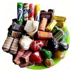 May include: A collection of assorted individually wrapped sweets and candies on a green plate. The items include cylindrical rolls with labels reading Herma, rectangular brown packages, and small, round, translucent treats in red, green, and brown.