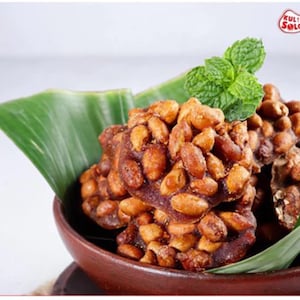 May include: A brown ceramic bowl filled with clusters of roasted peanuts coated in a dark, caramelised glaze. The snack is garnished with a sprig of fresh green mint leaves and rests against a large, vibrant green banana leaf. Text reads KULINER SOLOO.