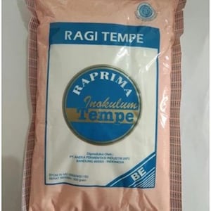 Op de afbeelding: Een rechthoekig plastic zakje met Raprima Ragi Tempe, een tempeh-startercultuur. De verpakking is licht perzikkleurig met een blauw centraal label met de tekst RAGI TEMPE, RAPRIMA, Inokulum Tempe en productiedetails inclusief 500 gram nettogewicht.