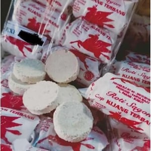 Può includere: Diversi biscotti rotondi, color bianco sporco e testurizzati, sono mostrati accanto a diverse confezioni di plastica trasparente. Ogni confezione presenta carta bianca con testo rosso Roti Sagon Cap Kijang Terbang e un grafico rosso di un cervo alato.
