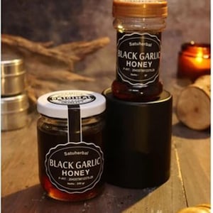 Puede incluir: Dos frascos de vidrio de miel de ajo negro Satuherbal. Un frasco con tapa blanca se asienta sobre la superficie, mientras que el otro con tapa dorada descansa sobre un soporte cilíndrico negro. Ambos frascos tienen etiquetas negras con texto blanco.