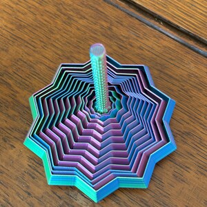 May include: A multi-layered, star-shaped top with a central post. The top features a gradient of colors, including blue, purple, and green. The design has a geometric, layered appearance, and the post is a metallic silver color.