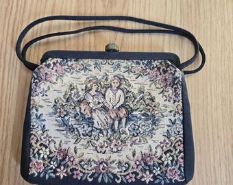 May include: A vintage tapestry handbag with a dark blue frame and handle. The bag has a clasp closure and a tapestry design featuring two figures in a garden scene. The floral tapestry includes pink, blue, and green hues.