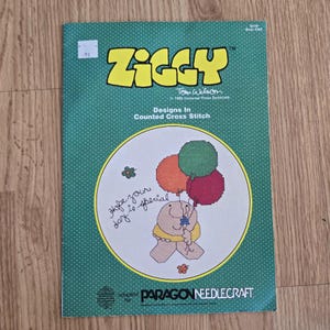 May include: A vintage "Ziggy" counted cross stitch pattern book. The cover is green with white polka dots and features a cross-stitched illustration of Ziggy holding balloons. Text includes "Hope your day is special" and "Paragon Needlecraft."