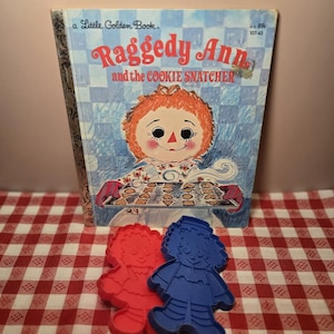 May include: A vintage "Raggedy Ann and the Cookie Snatcher" Little Golden Book with a red and blue Raggedy Ann cookie cutter set. The book cover features a smiling Raggedy Ann character holding a tray of cookies. The cookie cutters are in the shape of Raggedy Ann.