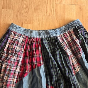 May include: A patchwork skirt featuring an elasticized waistband and a variety of fabric panels. The design incorporates red, white, and black plaid patterns, dark green and blue tartan sections, and solid light blue denim-like fabric segments.