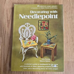 May include: The cover of a vintage book, "Decorating with Needlepoint", shows a white chair with a needlepoint cushion, a small table with a vase, and a framed needlepoint artwork. The cover is olive green with text and illustrations.