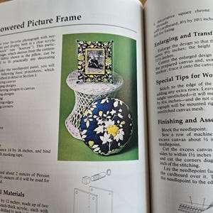 May include: A page from a craft book displays a framed picture on a white wicker stand, and a round cushion with a floral pattern. The frame has a black border with a floral design and a picture inside. The cushion is navy blue with white and yellow flowers.