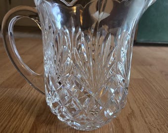 May include: A clear glass jug featuring a decorative cut pattern with a diamond-shaped base and vertical fan-like motifs extending towards the scalloped rim. The vessel includes a curved handle and a prominent pouring spout on one side.