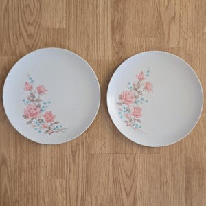 May include: Two white ceramic plates with a floral design. Each plate features a cluster of pink roses and small blue flowers on a stem. The plates are set against a light brown wooden surface.