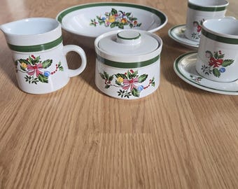 May include: A white ceramic tea set with a green trim and festive floral and bow designs. The set includes a creamer, sugar bowl, serving dish, and two teacups with saucers. The set is on a light brown wooden surface.