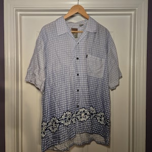 May include: Light blue short-sleeve shirt with a white dot pattern and a floral design at the bottom. The shirt has a button-down front, a chest pocket, and a collar. The shirt is hanging on a wooden hanger.
