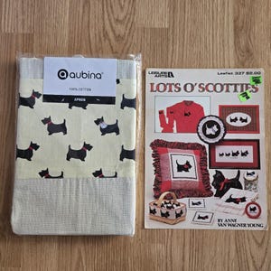May include: An apron with a repeating pattern of black Scottie dogs on a cream background, packaged with a craft pattern booklet titled "Lots O' Scotties." The apron is labeled "aubina" and made of 100% cotton.