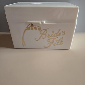 May include: A white rectangular box with a hinged lid. The box features gold lettering that reads "Bride's File" and a gold floral design. The box is likely for storing wedding-related items.