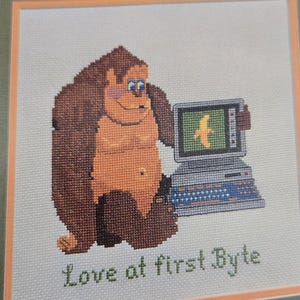 May include: Cross-stitch artwork featuring a cartoon gorilla holding a vintage computer with a banana icon on the screen. The text "Love at first Byte" is stitched below the image. The gorilla is brown and orange.