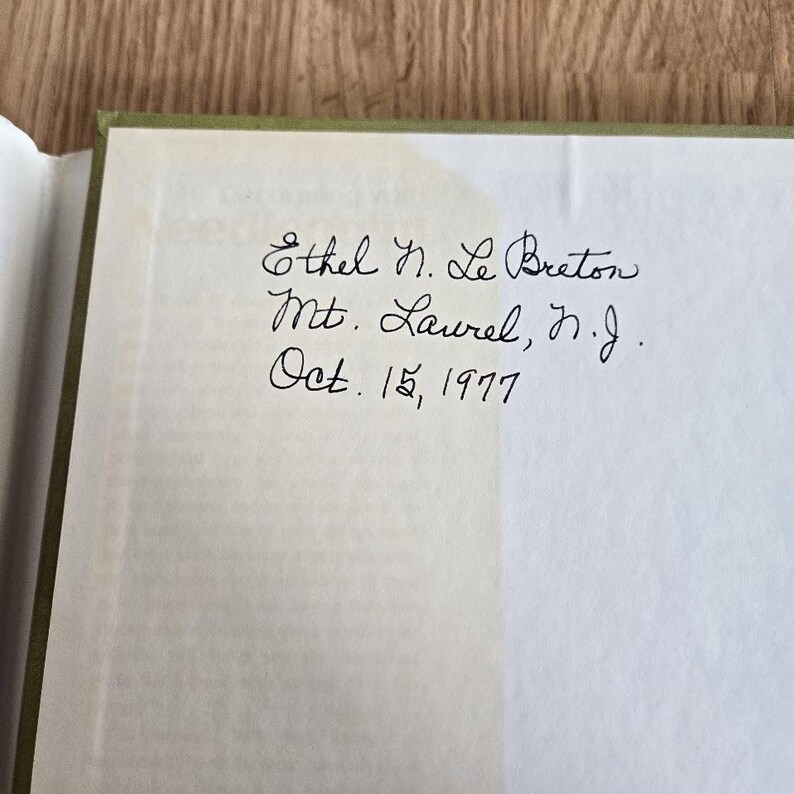 May include: An open book with a handwritten inscription in black ink. The text reads "Ethel N. Le Breton, Mt. Laurel, N.J., Oct. 15, 1977." The book's pages are off-white, and the binding is green.