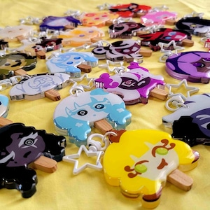 May include: A selection of colourful, popsicle-shaped keyrings with diverse character designs. Each keyring has a wooden stick and a star-shaped clasp. The keyrings are displayed on a yellow surface.