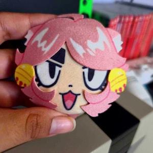 May include: A circular plush badge featuring a stylized anime-inspired character face with pink hair, large expressive eyes, a small mouth, and yellow circular accents on the sides. The design is printed on a soft, flat, felt-like material.