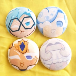 Pode incluir: Quatro pins redondos de personagens coloridos. Cada pin apresenta um personagem de desenho animado diferente com esquemas de cores azul, branco e bege. Os pins estão dispostos sobre um fundo amarelo brilhante.