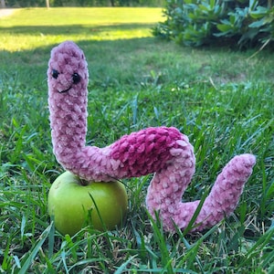 May include: A pink and purple crocheted worm toy with a smiling face is sitting on a green apple. The worm is on a bed of green grass.