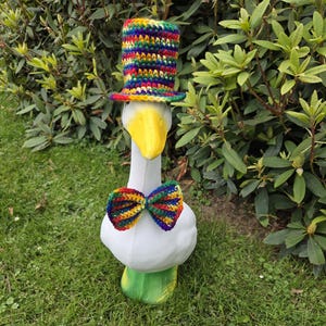 May include: A whimsical goose statue with a yellow beak, white body, and green base. It is adorned with a colorful, crocheted top hat and a matching bow tie, creating a playful and eye-catching garden decoration.