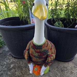 May include: A decorative white duck statue with a yellow beak and black eyes. The duck wears a colorful crocheted sweater and holds an orange pumpkin. The statue is placed in front of two black plant pots.