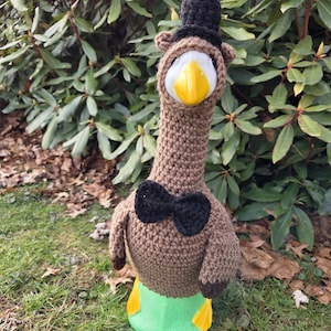 May include: A handmade crochet goose doll with a black top hat and bow tie. The goose has a white head with a yellow beak, brown body, and green base with yellow feet. The doll is standing on green grass.