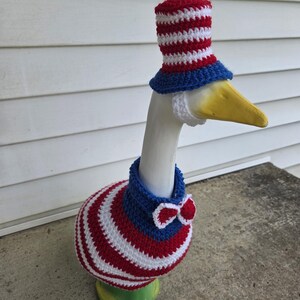 May include: A ceramic goose statue adorned with a patriotic crochet outfit. The outfit includes a red, white, and blue top hat, a blue collar with a red and white bow tie, and a body wrap with red and white stripes. The goose has a yellow beak.