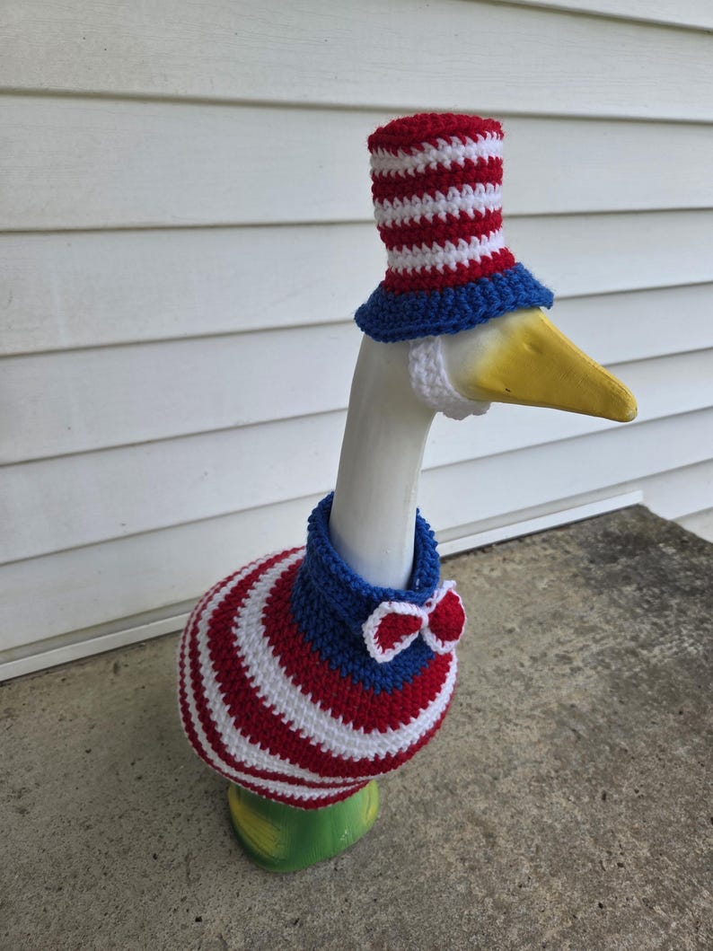 May include: A ceramic goose statue adorned with a patriotic crochet outfit. The outfit includes a red, white, and blue top hat, a blue collar with a red and white bow tie, and a body wrap with red and white stripes. The goose has a yellow beak.