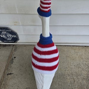 May include: A white goose wearing a patriotic crochet outfit. The outfit includes a red, white, and blue striped hat, a blue neck piece, and a red and white striped body cover. The goose is standing on a concrete surface.