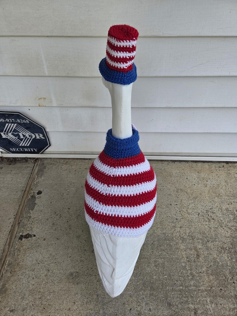 May include: A white goose wearing a patriotic crochet outfit. The outfit includes a red, white, and blue striped hat, a blue neck piece, and a red and white striped body cover. The goose is standing on a concrete surface.