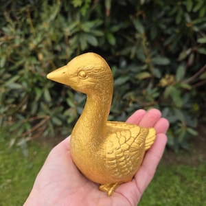 K&ouml;nnte beinhalten: Eine goldene Entenfigur mit detaillierten Federstrukturen. Die Ente befindet sich in einer sitzenden Position, mit erhobenem Kopf. Die Figur wird in einer Hand vor einem Hintergrund aus gr&uuml;nem Laub gehalten. Schnabel und F&uuml;&szlig;e der Ente sind ebenfalls golden.