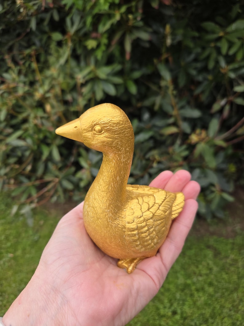 K&ouml;nnte beinhalten: Eine goldene Entenfigur mit detaillierten Federstrukturen. Die Ente befindet sich in einer sitzenden Position, mit erhobenem Kopf. Die Figur wird in einer Hand vor einem Hintergrund aus gr&uuml;nem Laub gehalten. Schnabel und F&uuml;&szlig;e der Ente sind ebenfalls golden.