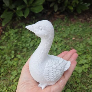 K&ouml;nnte beinhalten: Eine kleine, wei&szlig;e Keramik-Entenfigur mit detaillierten Feder- und Schnabelmerkmalen. Die Ente wird in einer Hand vor einem Hintergrund aus gr&uuml;nem Laub gehalten. Die Figur ist ein dekorativer Gegenstand.