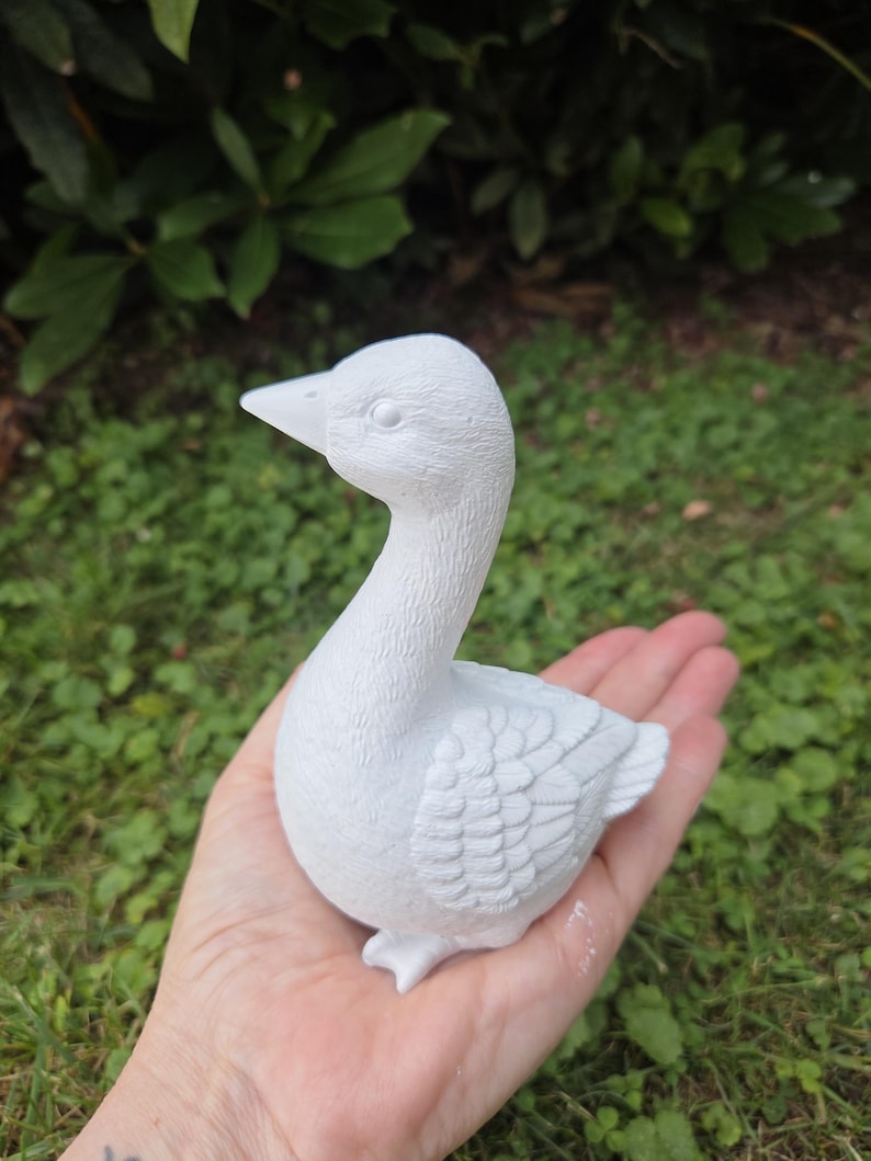 K&ouml;nnte beinhalten: Eine kleine, wei&szlig;e Keramik-Entenfigur mit detaillierten Feder- und Schnabelmerkmalen. Die Ente wird in einer Hand vor einem Hintergrund aus gr&uuml;nem Laub gehalten. Die Figur ist ein dekorativer Gegenstand.