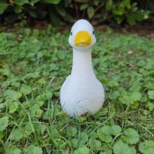 K&ouml;nnte beinhalten: Eine wei&szlig;e Entenfigur mit gelbem Schnabel und schwarzen Augen, platziert in Gras und Klee. Das Gartenornament hat eine strukturierte Oberfl&auml;che. Ein dekoratives Element f&uuml;r den Garten mit freundlichem Aussehen.