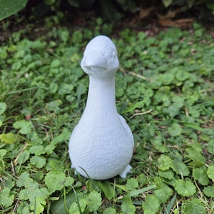K&ouml;nnte beinhalten: Eine kleine, wei&szlig;e G&auml;nsefigur steht auf einem Bett aus gr&uuml;nem Gras und Klee. Die Gans hat eine glatte, matte Oberfl&auml;che und ist aufrecht positioniert, mit Kopf und Schnabel nach vorne. Der Hintergrund zeigt &uuml;ppiges Gr&uuml;n.