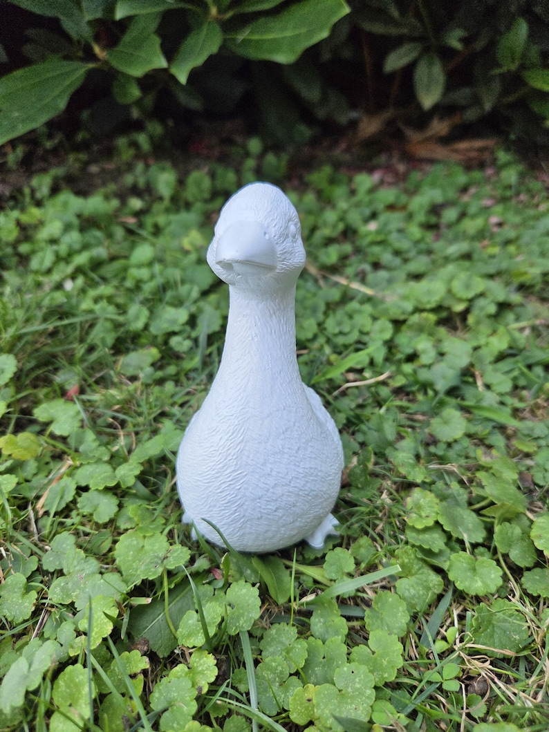 K&ouml;nnte beinhalten: Eine kleine, wei&szlig;e G&auml;nsefigur steht auf einem Bett aus gr&uuml;nem Gras und Klee. Die Gans hat eine glatte, matte Oberfl&auml;che und ist aufrecht positioniert, mit Kopf und Schnabel nach vorne. Der Hintergrund zeigt &uuml;ppiges Gr&uuml;n.