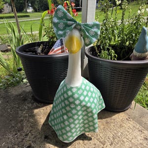 May include: A decorative goose statue with a white body, yellow beak, and a green bow and dress adorned with white floral patterns. The goose is positioned in front of two black flower pots, with an American flag visible in the background.