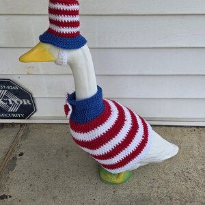 May include: A white goose statue wearing a patriotic crochet outfit. The outfit includes a red, white, and blue striped hat, a matching sweater, and a blue collar. The goose stands on a green base.