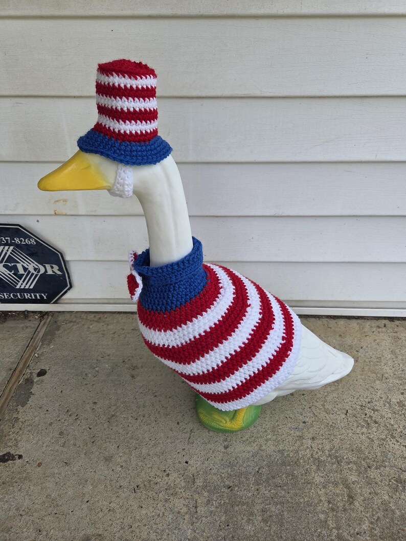 May include: A white goose statue wearing a patriotic crochet outfit. The outfit includes a red, white, and blue striped hat, a matching sweater, and a blue collar. The goose stands on a green base.