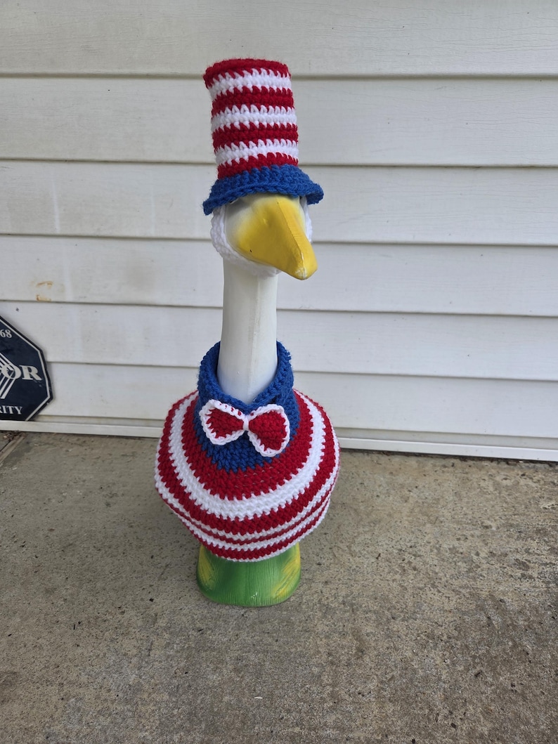 May include: A white goose wearing a red, white, and blue crocheted hat and a matching scarf with a blue bow tie.