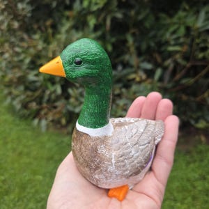 K&ouml;nnte beinhalten: Keramik-Entenfigur mit gr&uuml;nem Kopf, orangefarbenem Schnabel und F&uuml;&szlig;en sowie einem braunen und wei&szlig;en K&ouml;rper. Die Ente wird in einer Hand gehalten, wodurch ihre realistischen Merkmale und die strukturierte Oberfl&auml;che hervorgehoben werden. Dekorativer Artikel.