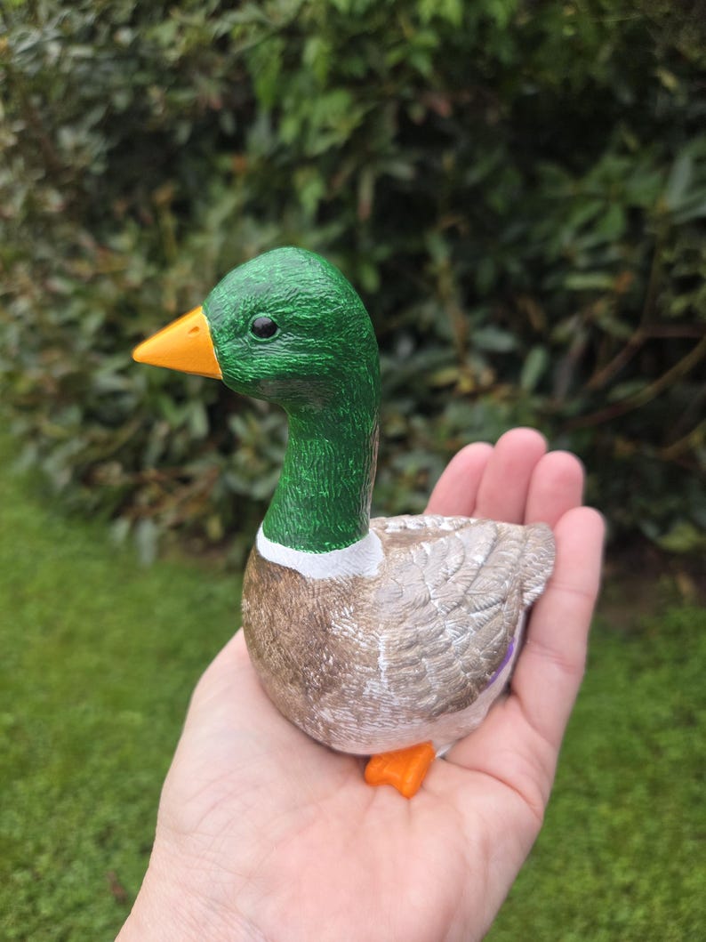 K&ouml;nnte beinhalten: Keramik-Entenfigur mit gr&uuml;nem Kopf, orangefarbenem Schnabel und F&uuml;&szlig;en sowie einem braunen und wei&szlig;en K&ouml;rper. Die Ente wird in einer Hand gehalten, wodurch ihre realistischen Merkmale und die strukturierte Oberfl&auml;che hervorgehoben werden. Dekorativer Artikel.