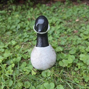K&ouml;nnte beinhalten: Eine Keramikgansfigur mit schwarzem Kopf und Hals, einem wei&szlig;en Wangenfleck und einem strukturierten, hellfarbigen K&ouml;rper. Die Gans befindet sich in der Mitte des Bildes, umgeben von gr&uuml;nem Gras und Bl&auml;ttern.