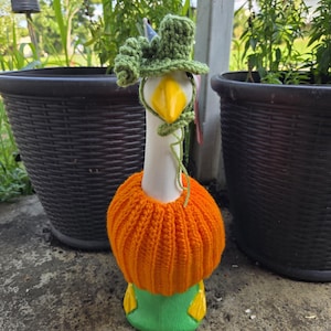 May include: A ceramic goose figurine with a yellow beak and green and orange knitted accessories. The goose wears a green hat and an orange knitted collar. The base of the goose is green with yellow accents. The background includes two black planters and greenery.