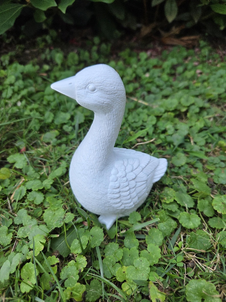 K&ouml;nnte beinhalten: Hellblaue G&auml;nsefigur mit detaillierten Federn und Augen. Die Gans steht auf gr&uuml;nem Gras und Klee. Die Figur hat einen gebogenen Hals und einen spitzen Schnabel, geeignet f&uuml;r Garten- oder Heimdekoration.