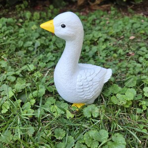 K&ouml;nnte beinhalten: Eine wei&szlig;e G&auml;nsefigur mit gelbem Schnabel und F&uuml;&szlig;en, vor gr&uuml;nem Gras und Laub. Die Gans hat eine detaillierte, strukturierte Oberfl&auml;che und ein schwarzes Auge, eine charmante Gartendekoration.